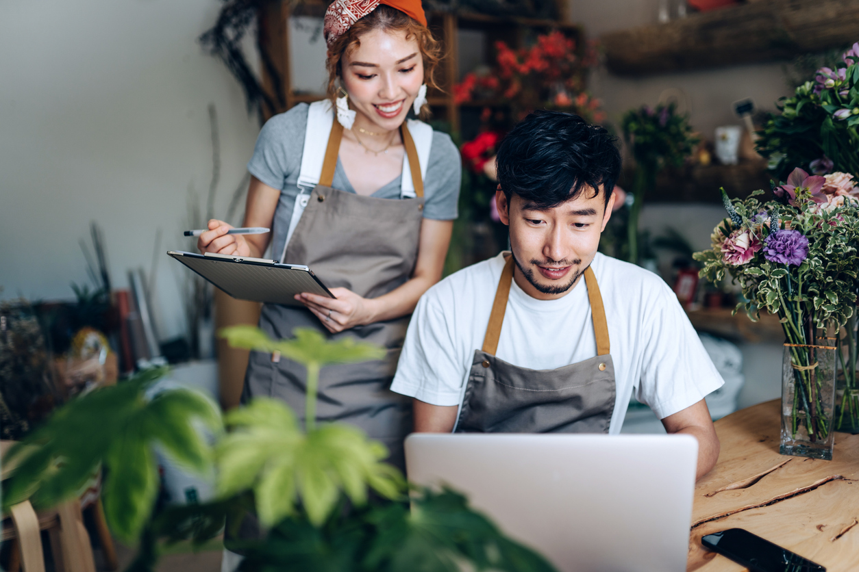 The young couple, who own a small flower shop, are working inside their shop. They are using computers and engaging in discussions about business planning as they search for a bank to partner with for their start-up business. Together, they are working diligently towards the success of their business.